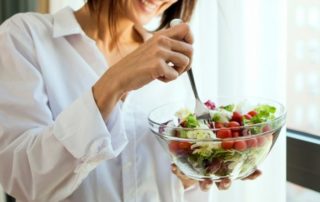 eine frau ist einen bunten salat aus einer gläsernen salatschüssel, die sie in ihrer linken hand hält