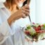eine frau ist einen bunten salat aus einer gläsernen salatschüssel, die sie in ihrer linken hand hält