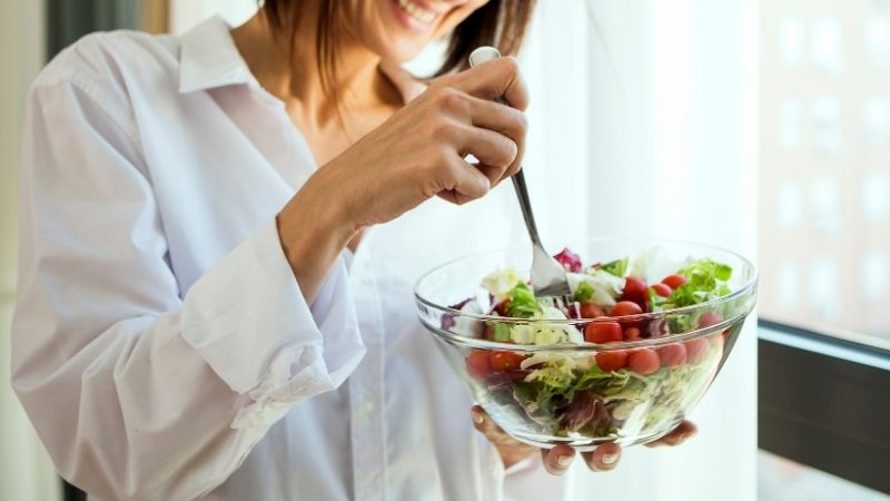eine frau ist einen bunten salat aus einer gläsernen salatschüssel, die sie in ihrer linken hand hält